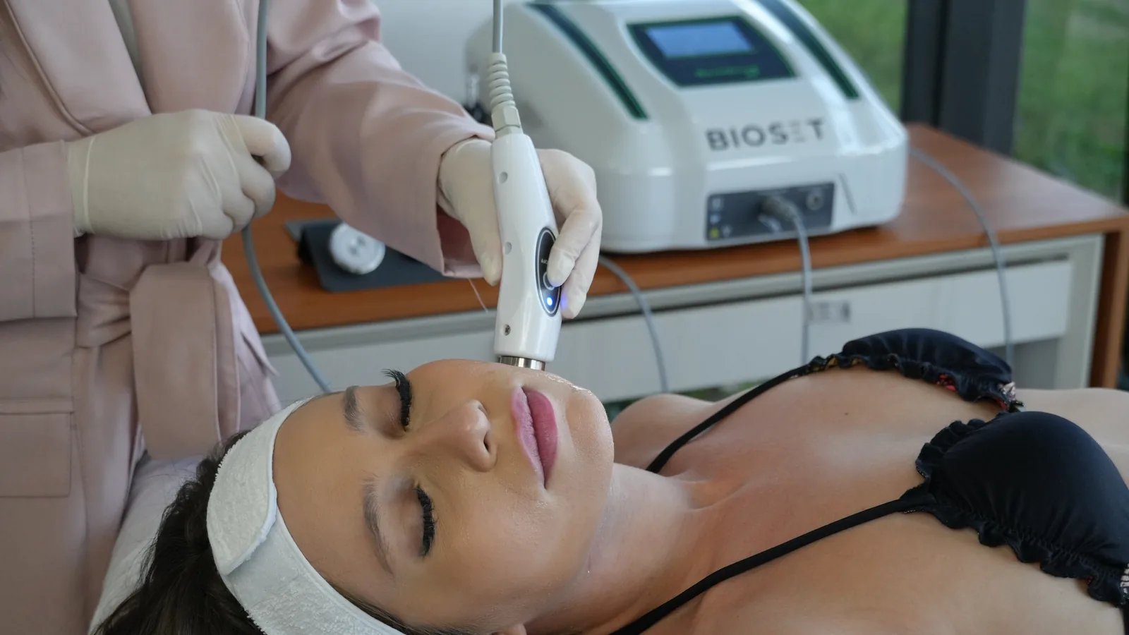 Profissional de luvas apoia o aplicador do Skinwave RF no queixo de uma paciente deitada, com o aparelho sobre a bancada ao fundo