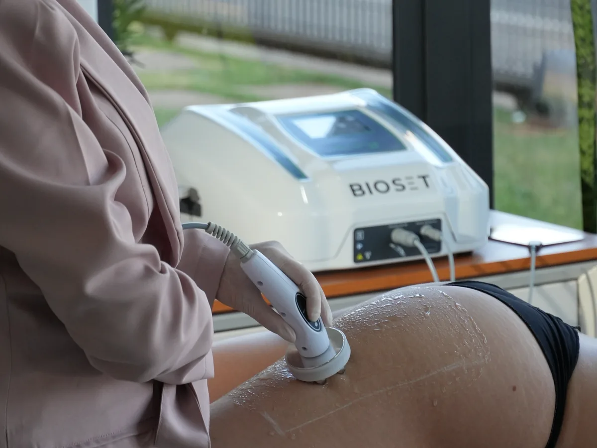 Profissional aplica o cabeçote do Skinwave RF na face posterior da coxa de paciente de bruços, com o aparelho na bancada ao fundo