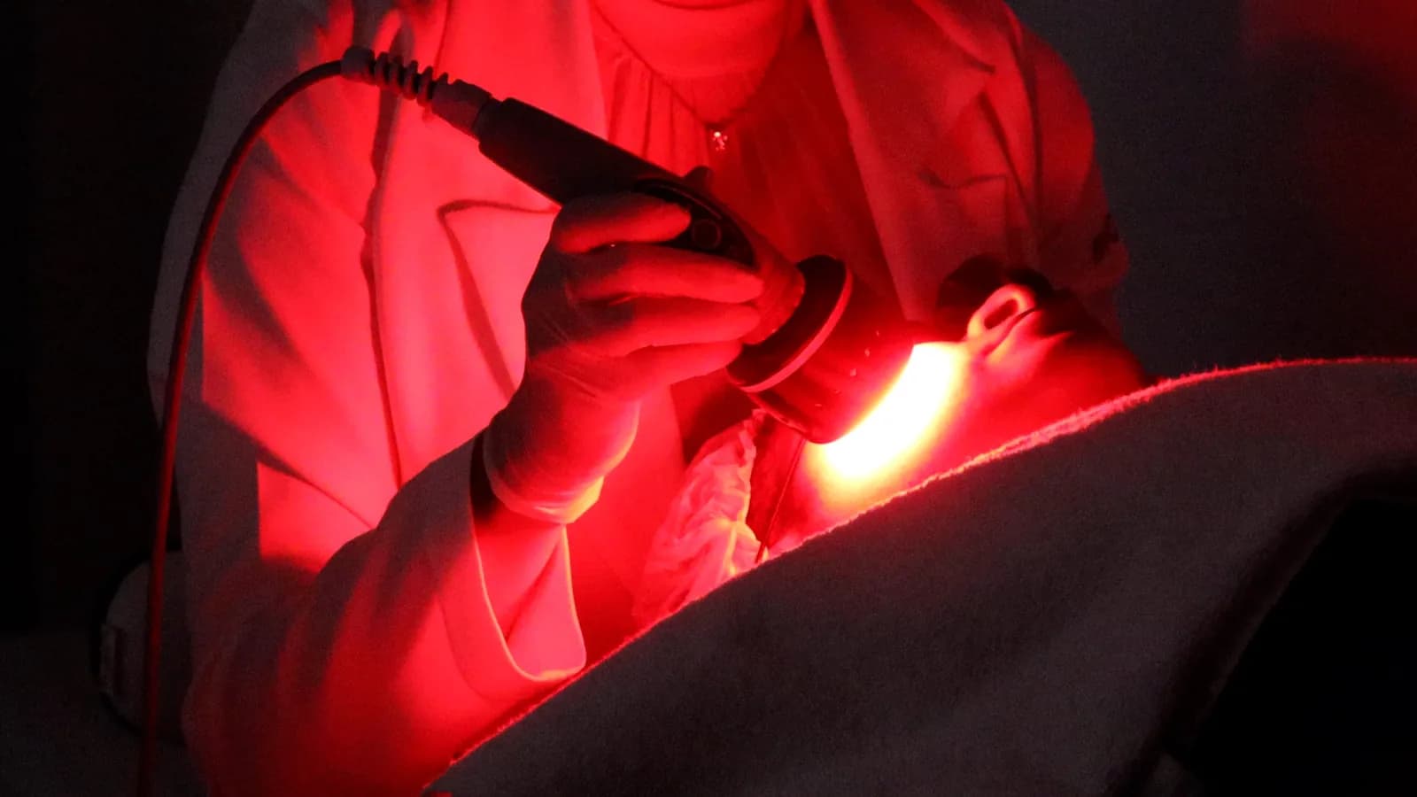 Profissional de jaleco e luvas aplica a ponteira de LED vermelho do Ledshape TT no rosto de uma paciente deitada e coberta por manta