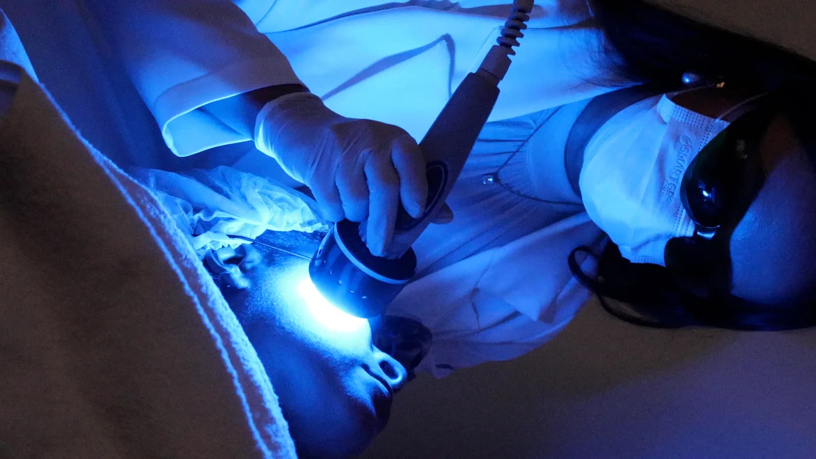 Profissional de máscara e óculos de proteção escuros aplica a ponteira de LED azul do Ledshape TT no rosto da paciente, vista de cima