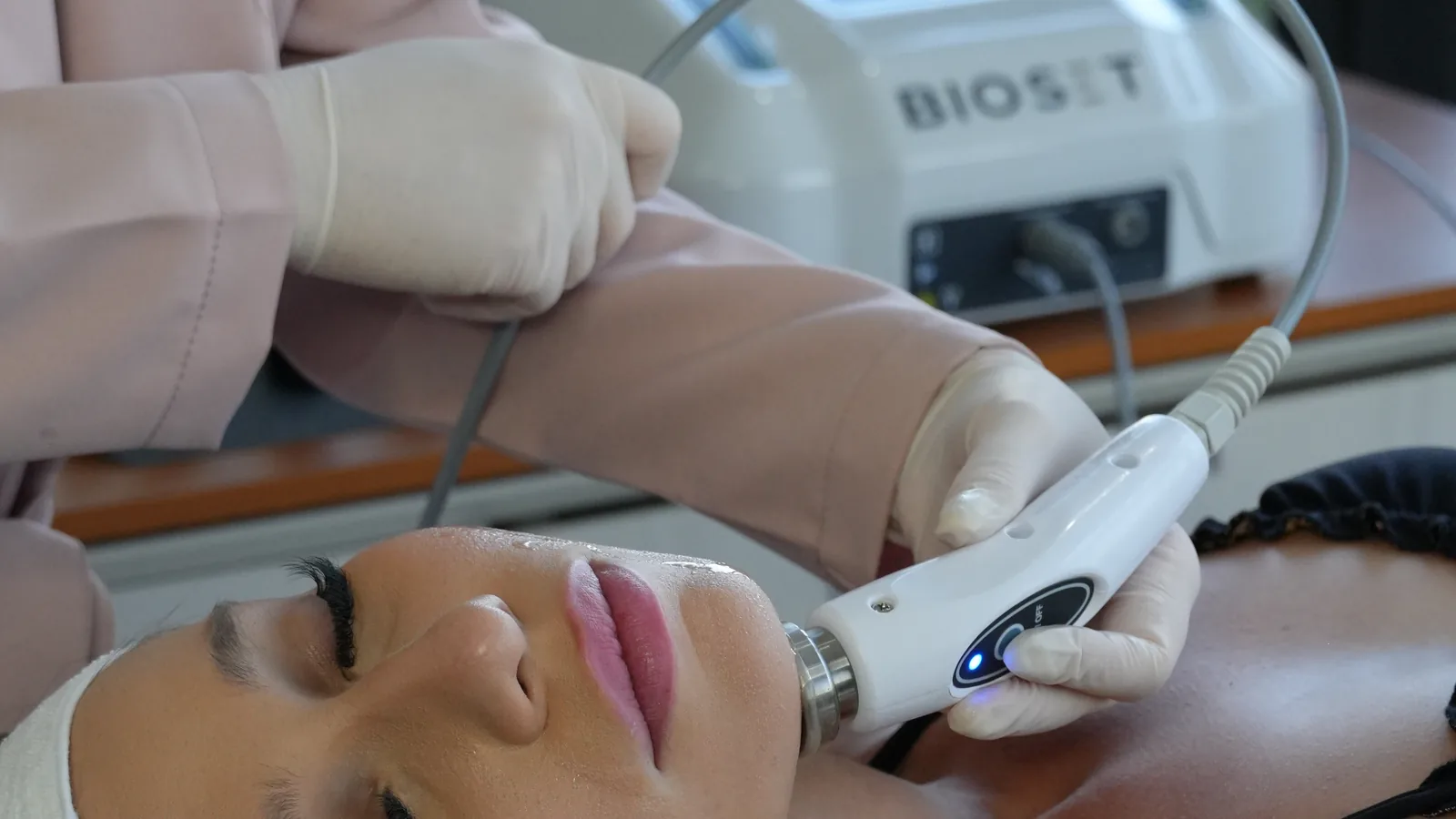 Mão enluvada conduz o aplicador do Skinwave RF sobre a mandíbula de uma paciente deitada, com o aparelho ao fundo