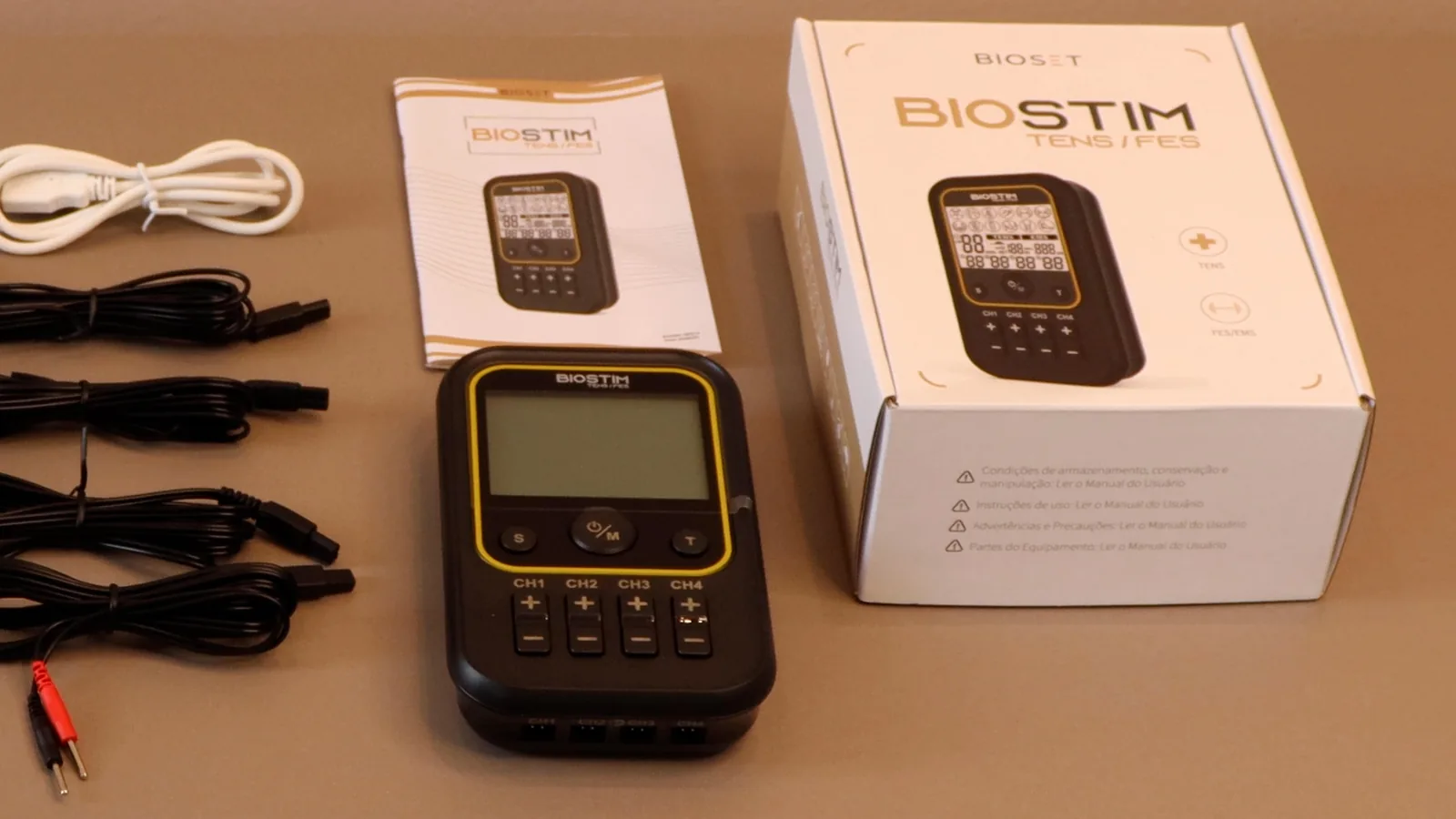 Biostim TENS e FES sobre a bancada, ao lado do manual do usuário e da caixa do produto, com cabos de eletrodo à esquerda